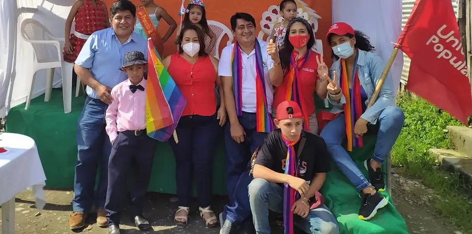 Grupo de personas posa bajo una carpa junto a banderas multicolores y rojas.