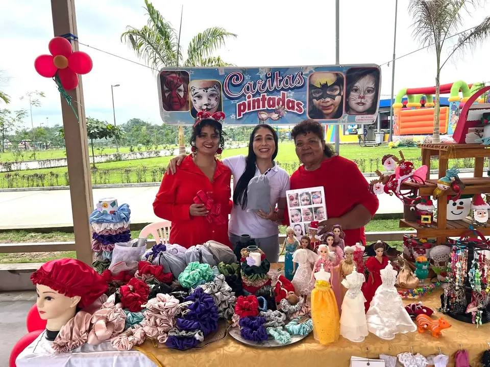 La autora posa entre dos emprendedoras frente a un puesto con accesorios, muñecas y otros productos.