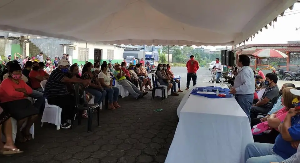 Público sentado bajo una carpa observa una mesa con micrófono y equipo de sonido.