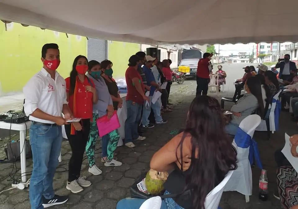 Personas sentadas y de pie participan en una reunión bajo una carpa; hay una mesa y altavoces.