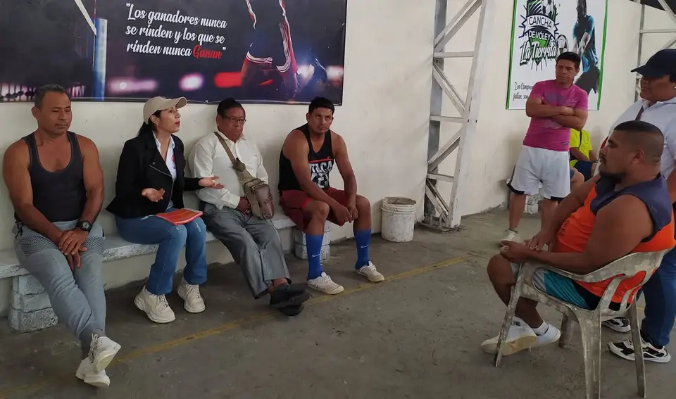 Personas conversan sentadas dentro de una cancha cubierta; hay carteles deportivos en la pared.