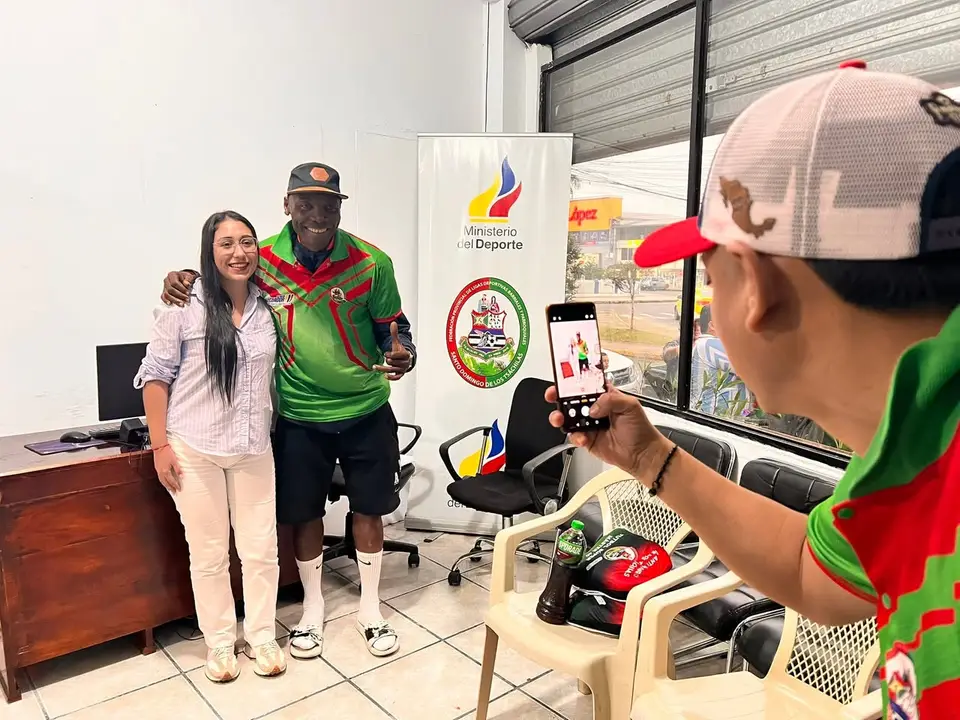 Mujer posa junto a un deportista uniformado dentro de una oficina; aparece un panel del Ministerio del Deporte.