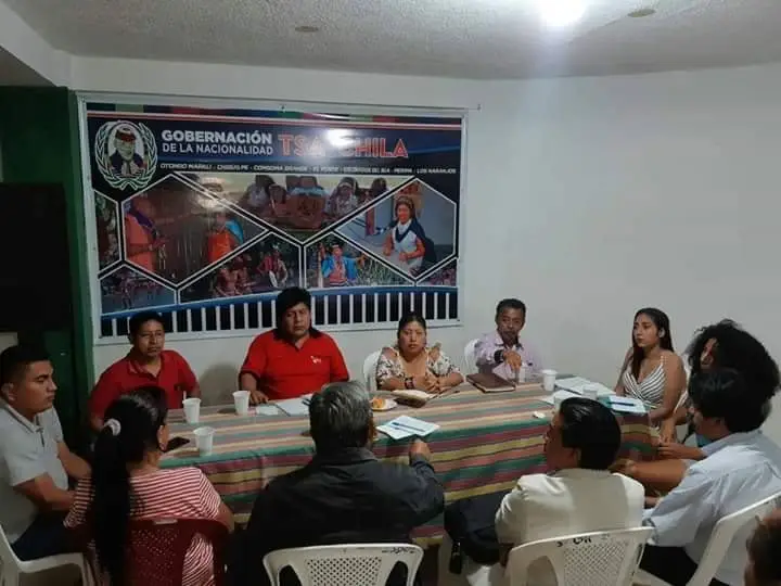 Varias personas están sentadas alrededor de una mesa con mantel a rayas; al fondo hay un cartel de la Gobernación de la Nacionalidad Tsáchila.