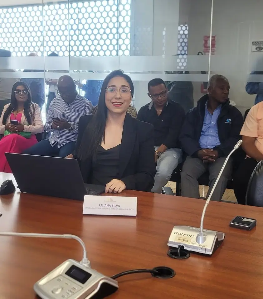 La autora participa en una reunión ante una mesa con micrófonos y una placa con su nombre.