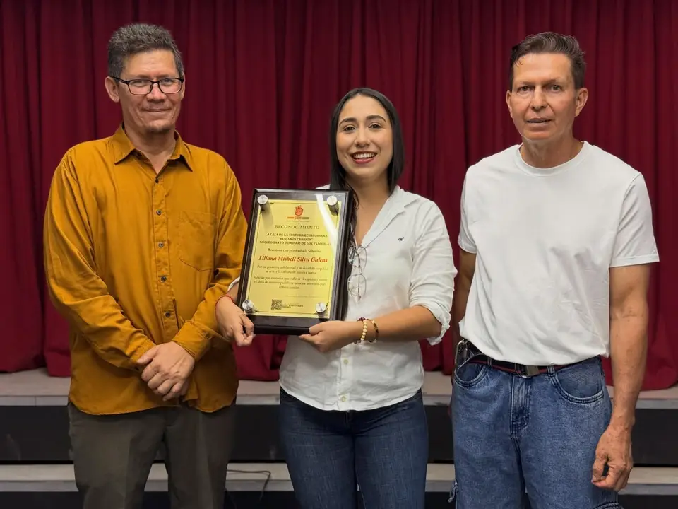 La autora sostiene una placa de reconocimiento junto a dos hombres ante un telón.