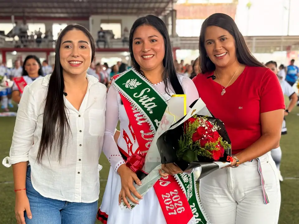 Tres mujeres posan en un recinto deportivo; la mujer central lleva una banda y sostiene un ramo de flores.