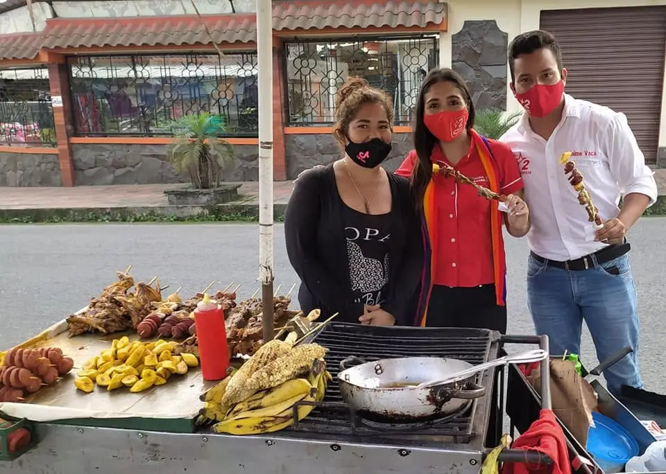 Tres personas posan detrás de un puesto de comida callejera con brochetas, plátanos y maíz.