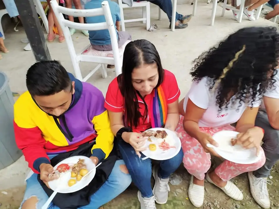 Tres personas comen de platos sentadas en sillas plásticas bajo techo.