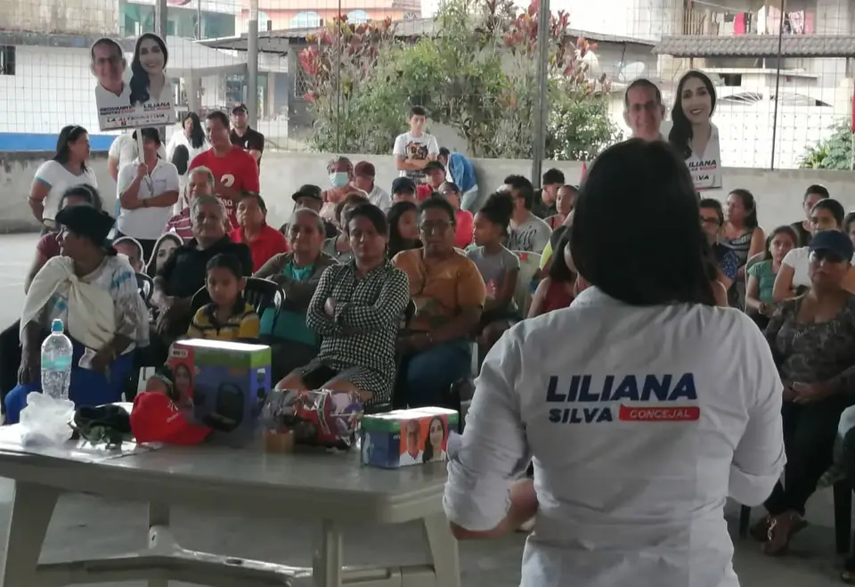 Reunión en una cancha cubierta; una mujer aparece de espaldas frente a personas sentadas.