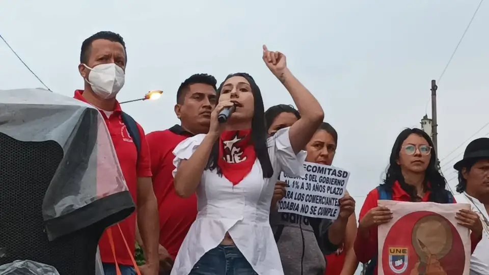 Persona habla con un micrófono y levanta el brazo ante varias personas y pancartas.