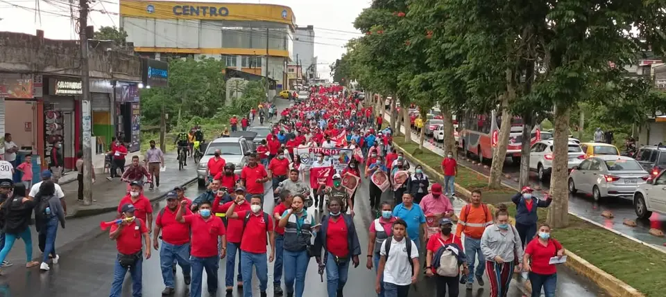 Manifestación multitudinaria avanza por una calle; predominan camisetas rojas y pancartas.