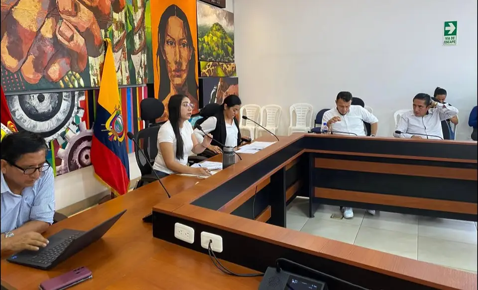 La autora participa en una sesión de comisión municipal ante una mesa con micrófonos; se observa la bandera de Ecuador.
