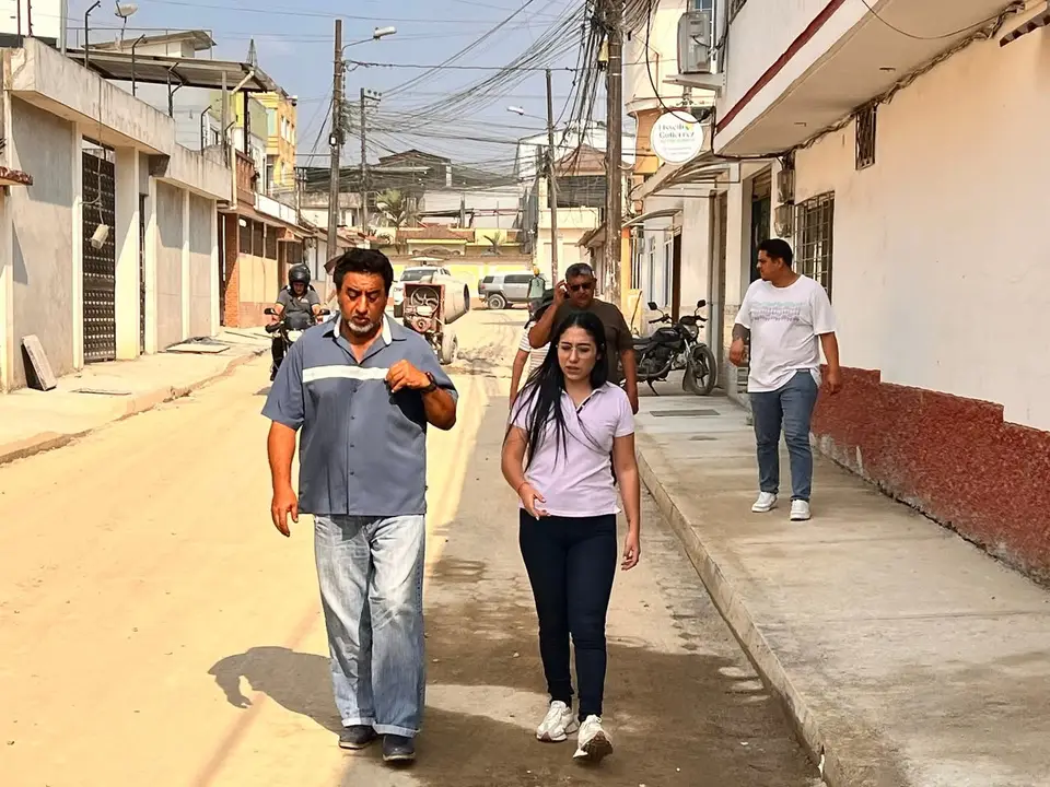 Mujer y hombre caminan por una calle urbana con viviendas, motocicletas y cableado aéreo.
