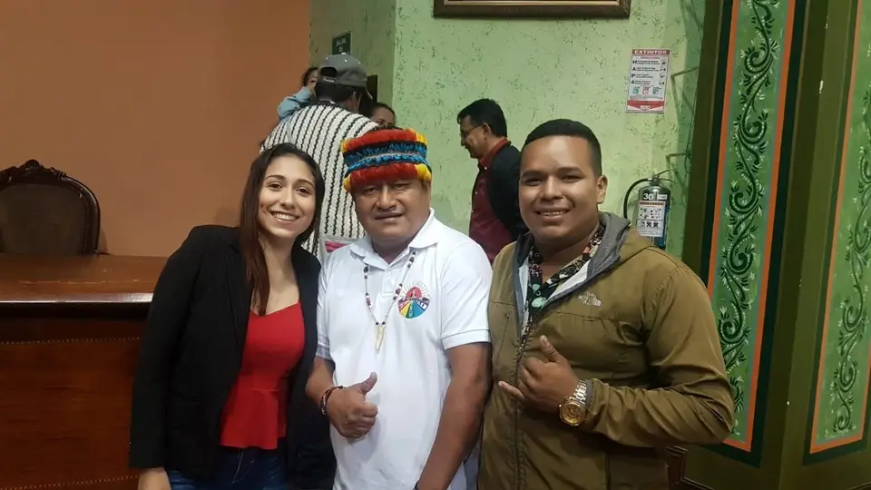 Tres personas posan en un salón; la persona del centro lleva un sombrero multicolor.