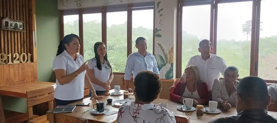 Personas participan en una reunión alrededor de mesas; una mujer habla de pie.