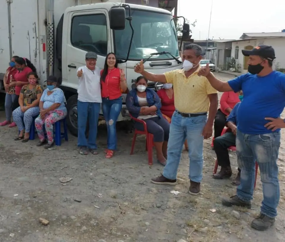 Grupo de personas se reúne al aire libre junto a un camión; varias están sentadas y otras levantan las manos.